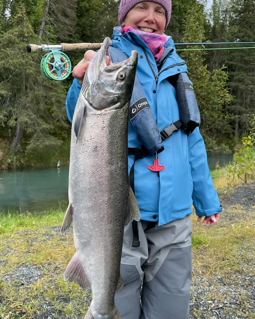 A person stands beside the Kenai River, holding up a large salmon they've caught