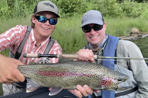 Two men posing with a large rainbow trout they caught.