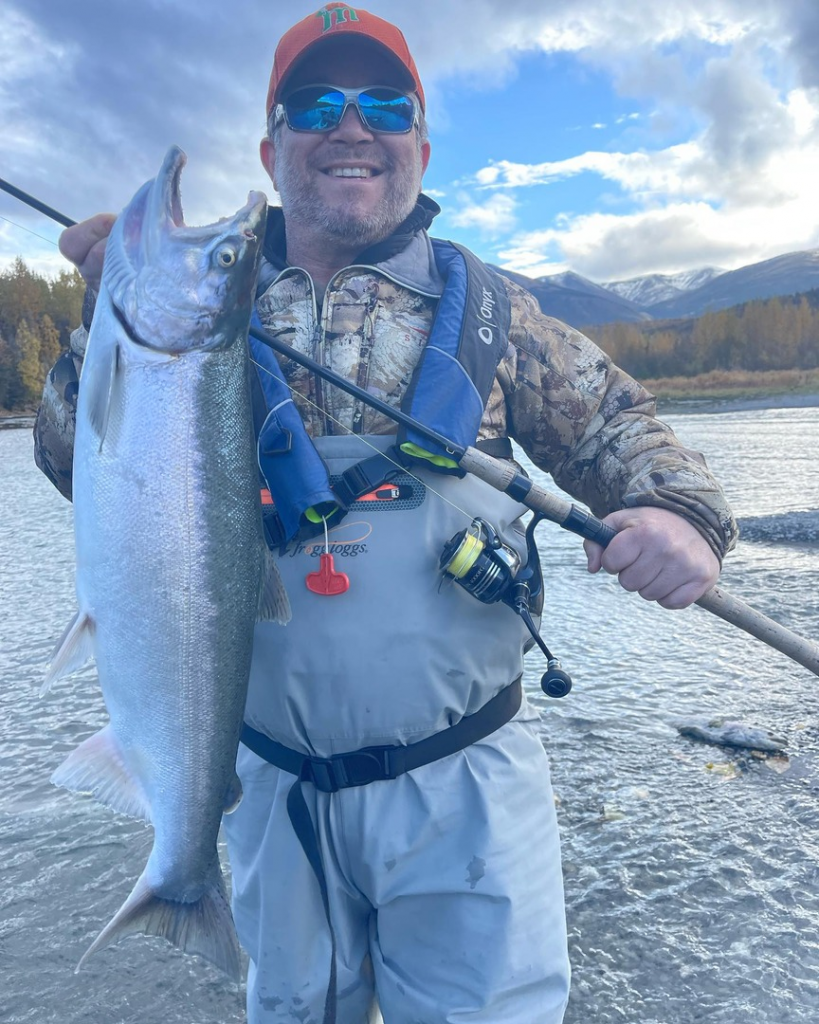 An angler proudly holding a large silver salmon caught from the Kenai River, with a backdrop of water and trees.