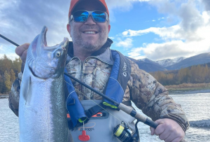 An angler proudly holding a large silver salmon caught from the Kenai River, with a backdrop of water and trees.