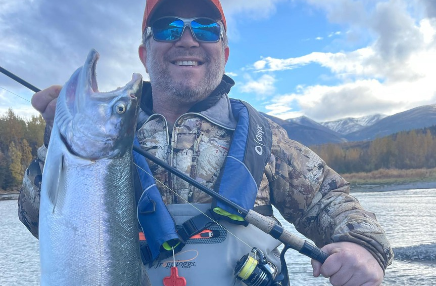 An angler proudly holding a large silver salmon caught from the Kenai River, with a backdrop of water and trees.