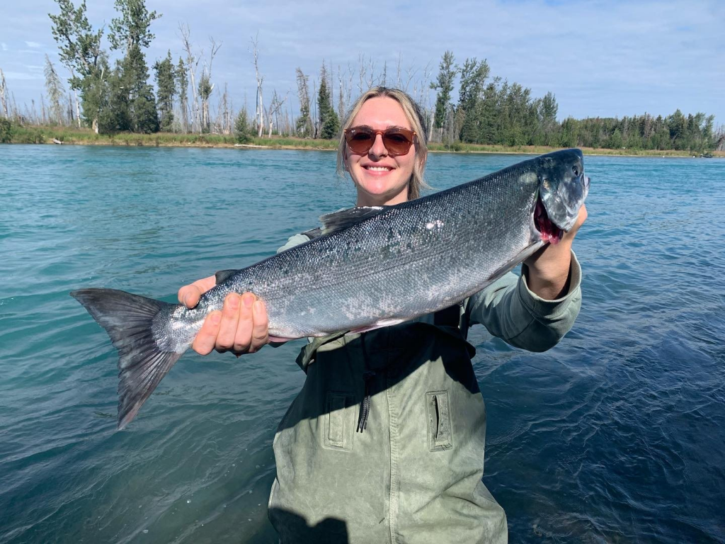 An angler standing in the clear waters of the Kenai River, holding a freshly caught silver salmon.