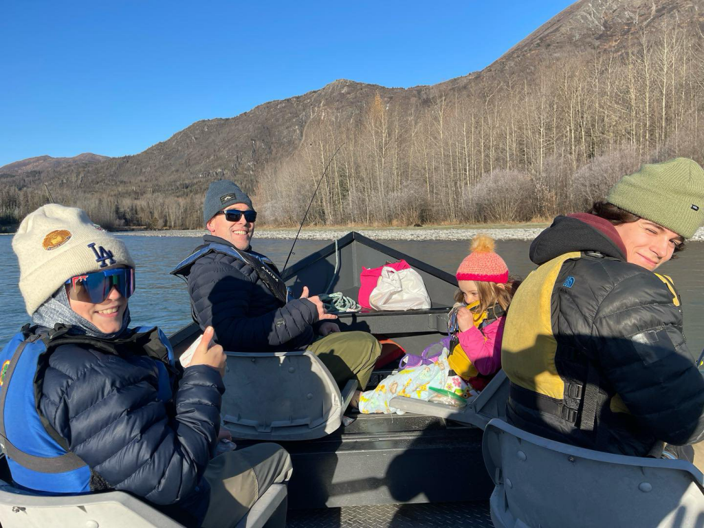 Three anglers and a child sitting in a drift boat on the Kenai River, enjoying a fishing trip.