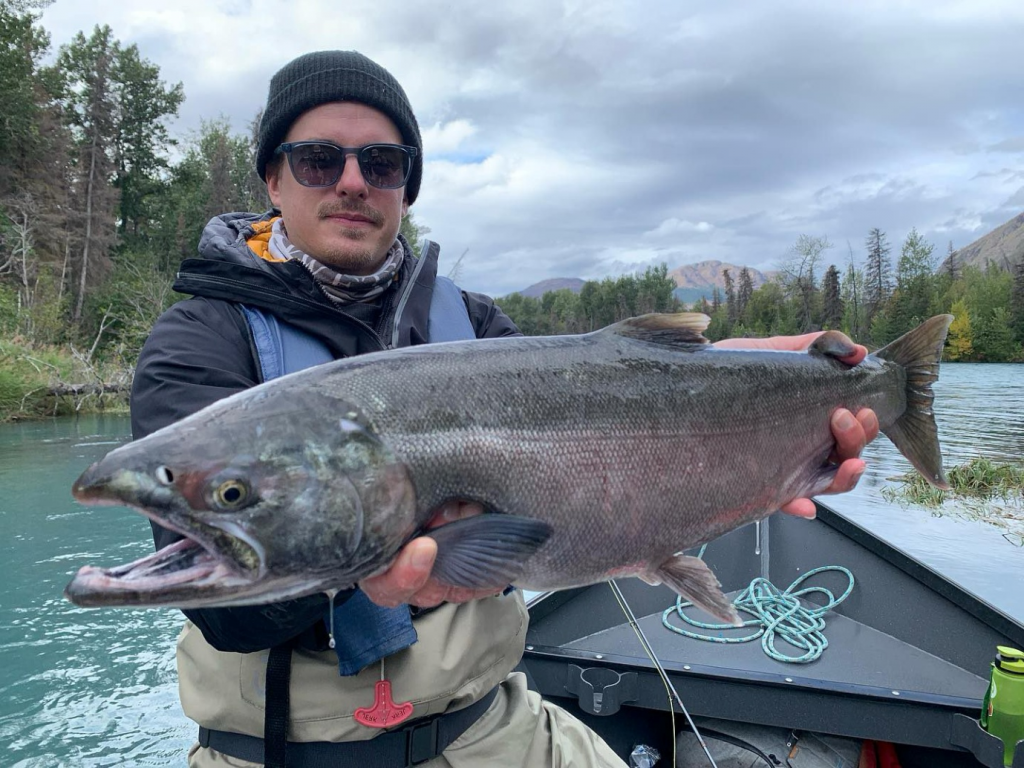 An angler on a drift boat in the Kenai River, holding a silver salmon with pride.